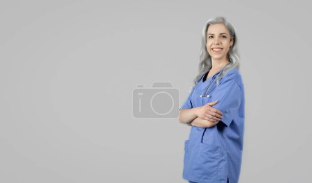 Téléchargez les photos : Offre soins de santé. Femme mûre souriante médecin en uniforme bleu posant avec stéthoscope regardant la caméra, debout près de l'espace de copie publicité clinique médicale sur fond de studio gris - en image libre de droit