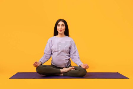 Photo for Pregnancy calm. Expecting woman in mindful meditation, sitting on yoga mat in lotus position and looking at camera over yellow background, free space - Royalty Free Image