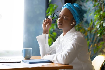Photo for Overworked tired young pretty african american woman in smart casual and traditional turban freelancer suffeing from headache, sitting at table in front of laptop, holding eyeglasses, sun flare - Royalty Free Image