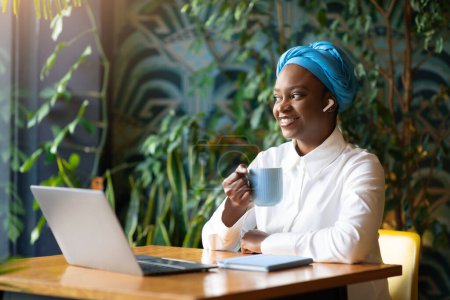 Téléchargez les photos : Heureuse jeune femme noire avec des écouteurs sans fil écouter de la musique sur ordinateur portable, boire du café tout en travaillant ou en étudiant au café, en utilisant la puissance des mélodies pour améliorer sa productivité, copier l'espace - en image libre de droit