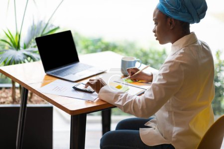 Photo for Young african american woman i smart casual working on budget, counting monthly expenses, sitting at cafe, using laptop with blank screen, calculator, taking notes, mockup, sun flare - Royalty Free Image