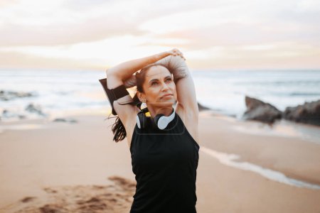 Photo for Positive mature european lady in sportswear and headphones doing stretching for arms, warming up, enjoy workout in morning on sea beach, outdoor. Fitness and body care, weight loss sport - Royalty Free Image