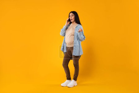 Photo for Young emotional pregnant woman talking on cellphone and gesturing, having phone conversation with lover or doctor while standing over yellow studio background, full length shot, free space - Royalty Free Image