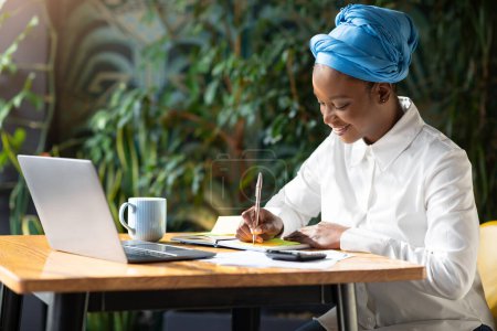Foto de Emprendimiento, negocios. Feliz joven mujer afroamericana emprendedora trabajando en su startup en la cafetería, sentada en la mesa frente a la computadora portátil, tomando notas en el bloc de notas, espacio para copiar - Imagen libre de derechos