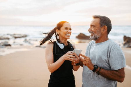 Photo for Smiling mature european woman and man in sportswear rest from workout together with smartphone in morning, have fun on sea beach, outdoor. Body care, sports app and blog, fitness and love - Royalty Free Image