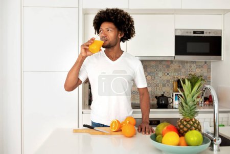 Téléchargez les photos : Rafraîchissements santé. Heureux homme afro-américain buvant du jus d'orange à partir de verre, jouissant d'un goût frais et hydratant debout dans la cuisine moderne intérieure. Hydratation et boissons Recette - en image libre de droit