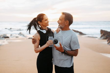 Photo for Cheerful mature european woman and man in sportswear rest from workout, drink bottle of water in morning on sea beach. Health care, sports, fitness together and body care outdoor - Royalty Free Image