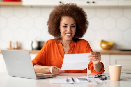 Happy Black Woman Reading Letter While Sitting At Desk With Laptop In Kitchen, Joyful African American Lady Emotionally Reacting To Good News In Mail, Got Promotion Or University Accceptance