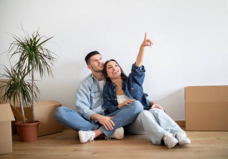 Photo for Dreamy young couple planning interior design after moving home, thinking about decorations and furniture in their new house, smiling european spouses sitting on floor and pointing up at copy space - Royalty Free Image