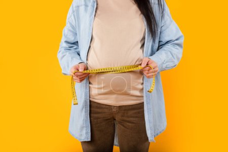 Photo for Mom-to-bes progress. Young pregnant woman measuring her belly with tape, standing on yellow background, cropped, closeup. Prenatal care, maternity, health, gynecology, medicine - Royalty Free Image