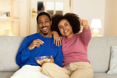 Téléchargez les photos : Le passe-temps domestique. heureux couple noir avec pop-corn regarder film à la télévision, femme pointant doigt assis à la maison. Aimer les jeunes conjoints relaxant reposant sur un canapé confortable le week-end - en image libre de droit