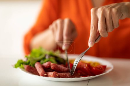 Photo for Unrecognizable african american woman eating tasty lunch in kitchen, having plate with fresh salad, pepperoni sausages and tomatoes, black lady enjoying meal, using fork and knife, closeup, cropped - Royalty Free Image