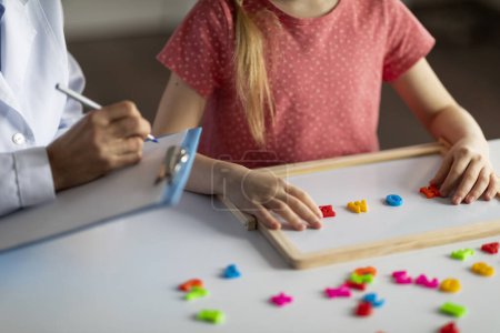 Photo for Unrecognizable little girl exercising with alphabet board during session with female child development specialist at clinic, small kid making words from colorful plastic letters, cropped image - Royalty Free Image