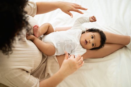 Photo for Happy black millennial mom playing with little baby in white clothes, lady enjoy motherhood in bedroom, top view. Child care vacation, parenthood and family at home, fun together - Royalty Free Image
