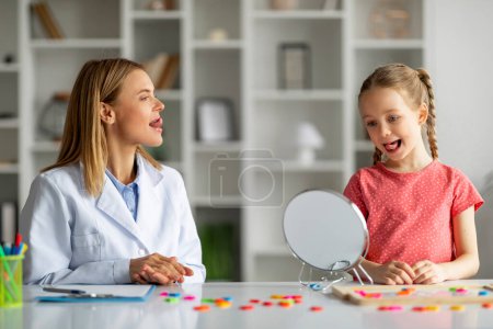 Photo for Kid with stuttering problem visiting speech-language therapist at clinic, cute little girl looking at mirror and showing tongue, repeating after female specialist, working on pronunciation - Royalty Free Image