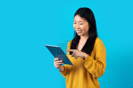 Foto de Feliz joven asiática atractiva dama en camisa amarilla disfrutando de su nueva tableta digital gadget, tocando la pantalla de la almohadilla, aislado en el fondo del estudio azul, desplazamiento, utilizando buena aplicación, espacio de copia para el anuncio - Imagen libre de derechos