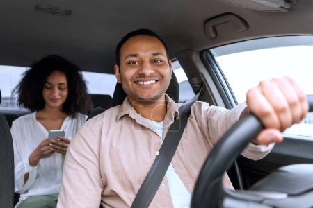 Photo for Car Ride. Cheerful Middle Eastern Driver Guy Posing At Wheel Driving Auto, While Passenger Woman Using Cellphone Application For Navigation And Communication Sitting On Vehicle Back Seat Inside - Royalty Free Image