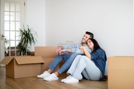 Photo for Dreamy Young Couple Sitting On Floor In Their New Apartment After Moving, Happy European Spouses Relaxing Among Cardboard Boxes With Belongings, Thinking About Home Interior Design, Copy Space - Royalty Free Image