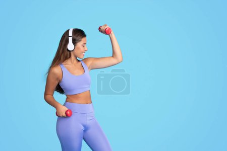 Photo for Happy young european slim woman athlete in sportswear and wireless headphones doing exercises with dumbbells, isolated on blue studio background. Sports, fit, body care and workout with music - Royalty Free Image