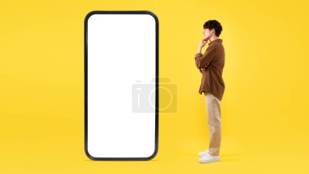 Photo for Mobile Offer. Thoughtful Guy Looking At Big Smartphone With Blank Screen, Thinking About Innovative Application Standing On Yellow Background. Man Posing Near Huge Gadget. Studio Shot, Panorama - Royalty Free Image