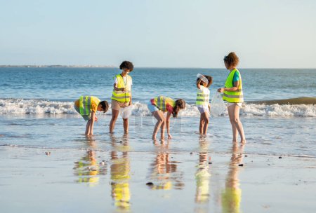 Téléchargez les photos : Heureux différents adolescents enfants bénévoles et enseignants en gilets collectent les ordures dans un sac pour le recyclage sur la plage de la mer, en plein air. Protection de l'environnement, éco nature, arrêt plastique et travail d'équipe - en image libre de droit