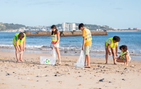 Téléchargez les photos : Heureux divers adolescents enfants bénévoles et femme dans des gilets collectent les ordures dans des sacs pour recyclage sur la plage, en plein air. Travail d'équipe, respect de l'environnement, éco nature, lutte contre le plastique - en image libre de droit