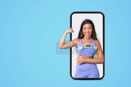 Photo for Cheerful young european slim lady in sportswear hold scales, show muscles biceps on screen big phone isolated on blue studio background. App for sports, fitness, body care blog, strength training - Royalty Free Image