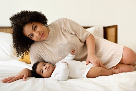 Foto de Feliz millennial negro más tamaño mamá juega con el pequeño bebé en la cama blanca, disfruta del amor y el tiempo libre en el dormitorio. Cuidado de niños, maternidad, paternidad y familia en el hogar, anuncios y ofertas - Imagen libre de derechos
