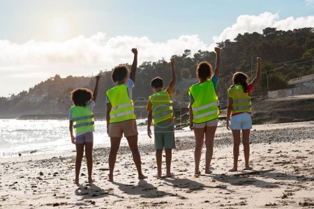 Photo for Happy confident diverse teen kids volunteers in vests raise hand up, celebrating victory over trash on beach, outdoor, back. Caring for environment, eco nature, fight against garbage and plastic - Royalty Free Image