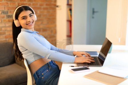 Téléchargez les photos : Adolescente indienne utilisant un ordinateur portable faisant ses devoirs en ligne et tapant sur le clavier, portant un casque et souriant à la caméra, assise à table à la maison, espace libre - en image libre de droit
