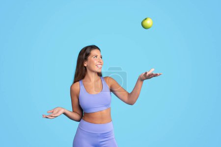 Photo for Smiling young european slim woman athlete brunette in sportswear tossing green apple, has fun isolated on blue studio background. Sports, fitness, body care and vitamins food - Royalty Free Image