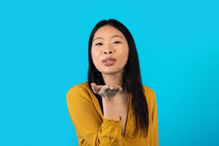 Photo for Beautiful cheerful young asian woman outstretching hand towards camera and showing fly kiss, chinese lady expressing love and affection on blue studio background, copy space - Royalty Free Image