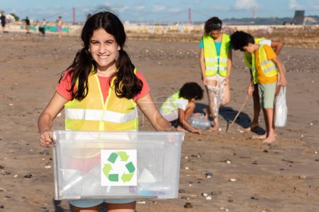 Foto de Sonriente chica árabe adolescente lleva caja para el reciclaje, niños multiétnicos voluntarios en chalecos recogen basura en la playa, al aire libre. El cuidado del medio ambiente, eco, lucha contra el plástico - Imagen libre de derechos