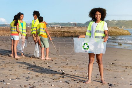 Téléchargez les photos : Heureux différents adolescents enfants bénévoles dans des gilets collectent les ordures, fille noire porte boîte pour le recyclage sur la plage, en plein air. Environnement de soins, éco nature, lutte avec le plastique - en image libre de droit