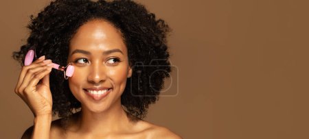 Téléchargez les photos : Joyeux sourire joyeux attrayant moitié nue jeune femme noire faisant massage du visage avec rouleau de jade dans la routine de soins du visage du matin, en regardant l'espace de copie, gros plan, fond de studio, bannière - en image libre de droit