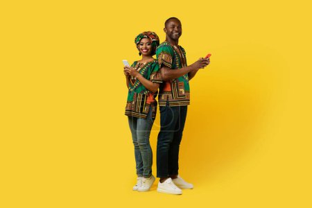 Photo for Happy black couple middle aged man and young woman in national african costumes using smartphones on yellow studio background, chatting, using dating app, full length, copy space - Royalty Free Image