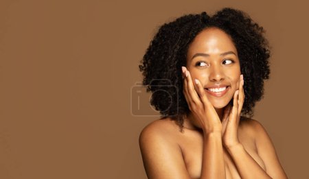 Foto de Rutina de cuidado de la piel no tóxica. Alegre desnudo bastante joven afroamericana mujer con el pelo tupido tocando su piel de la cara brillante, sonriendo y mirando el espacio de copia, fondo de estudio marrón, web-banner - Imagen libre de derechos