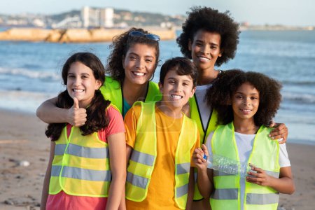 Téléchargez les photos : Des adolescentes internationales souriantes bénévoles et enseignantes dans des gilets pare-balles, montrent la bouteille et le pouce sur la plage, en plein air. Protection de l'environnement, éco nature, lutte contre le plastique, déchets - en image libre de droit