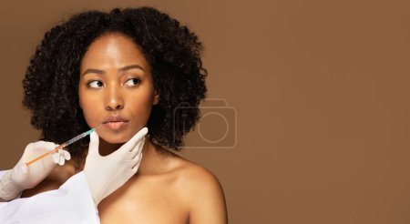 Photo for Pros and cons of lip fillers. Attractive half-naked young black woman getting lip injection on brown studio background, looking at copy space, beautician hands holding syringe, web-banner - Royalty Free Image