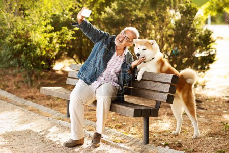 Téléchargez les photos : Heureux vieil homme européen décontracté aime marcher avec le chien, s'asseoir sur le banc dans le parc, prendre des photos sur smartphone en plein air. Selfie avec animal de compagnie, amour, ami, soins et temps libre et appel vidéo - en image libre de droit