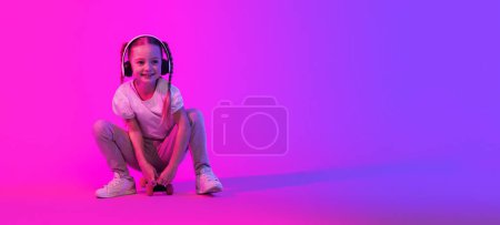 Téléchargez les photos : Adorable joyeux jolie fille d'âge scolaire en tenue d'été assis sur planche à roulettes sur fond futuriste, regardant l'espace de copie et souriant, en utilisant des écouteurs sans fil, écouter de la musique, bannière web - en image libre de droit
