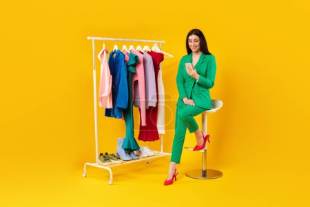 Photo for Trendy young shopaholic lady with cellphone shopping online while sitting near clothing rail, choosing new summer outfit and consulting with stylist, yellow background, full length - Royalty Free Image
