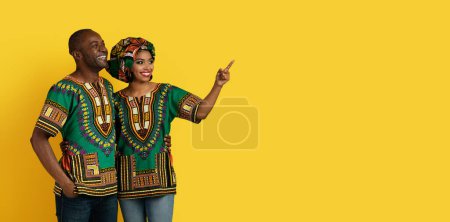 Photo for Great offer, deal concept. Loving cheerful beautiful black lovers wearing african national costumes embracing, pointing at copy space for advertisement on yellow studio background, panorama - Royalty Free Image