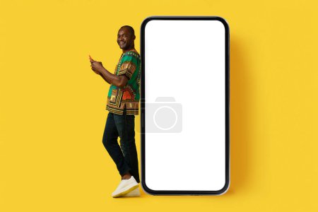 Photo for Happy cheerful handsome middle aged black man wearing national african shirt using nice mobile app on yellow background, standing by huge smartphone with white blank screen, mockup - Royalty Free Image