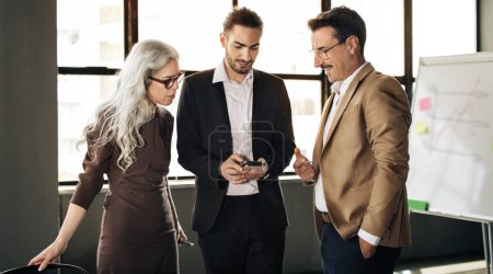 Photo for Corporate Meeting. Three Successful Middle Eastern Colleagues Using Business App On Smartphone, Standing Near Whiteboard In Office. Company Managers Trio Websurfing Via Phone At Workplace. Panorama - Royalty Free Image