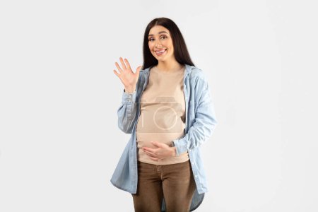 Photo for Positive young expectant woman touching belly and waving at camera saying hello and smiling, posing on white studio background. Friendly welcome gesture - Royalty Free Image