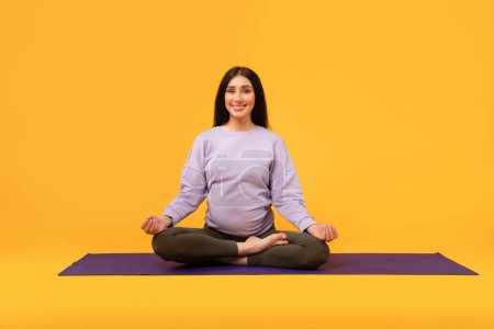 Téléchargez les photos : La force tranquille de la maternité. Heureuse femme enceinte assise sur un tapis de yoga, méditant et souriant, faisant de l'exercice sur fond jaune, copiant l'espace - en image libre de droit