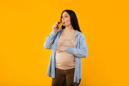 Foto de Mujer embarazada joven con gran barriga bebiendo un vaso de agua, disfrutando de la futura maternidad, de pie sobre un fondo amarillo. Equilibrio acuático, cuidado de la salud, expectativa del niño - Imagen libre de derechos