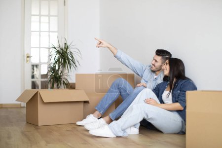 Photo for Design Ideas. Happy Young Couple Sitting On Floor Among Cardboard Boxes In New Flat And Pointing Away, Smiling European Spouses Thinking About Decorations And Furniture After Moving Home, Copy Space - Royalty Free Image