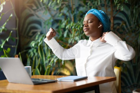 Photo for Exhausted unhappy young black businesswoman working online, sitting at table next to window at cafe, using pc laptop and earpods, stretching body with closed eyes, have difficulties in business - Royalty Free Image